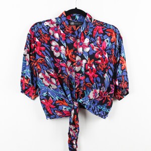 May include: A cropped, short-sleeved button-down shirt with a tie front. The shirt is made of a silky fabric and features a vibrant floral print in shades of pink, red, blue, and green on a black background. The shirt has a collar and a button closure. The label "AIM" is visible on the collar.