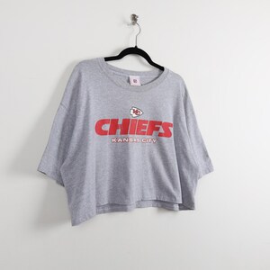 May include: A gray Chiefs t-shirt with a red logo and the words "CHIEFS" and "KANSAS CITY" printed on the front. The shirt is hanging on a black hanger against a white background.