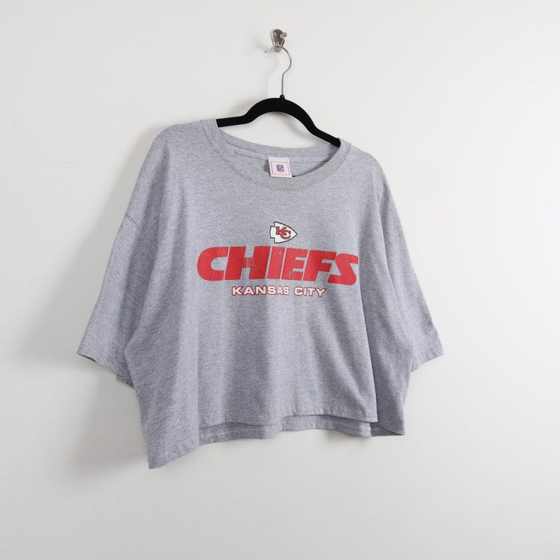 May include: A gray Chiefs t-shirt with a red logo and the words "CHIEFS" and "KANSAS CITY" printed on the front. The shirt is hanging on a black hanger against a white background.