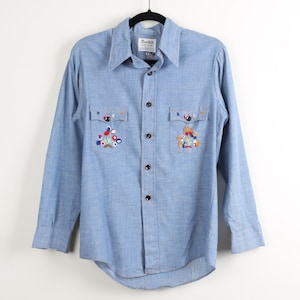 May include: Light blue button-down shirt with long sleeves and two chest pockets. The shirt features colorful embroidered floral designs on the pockets. The shirt is hanging on a black hanger.