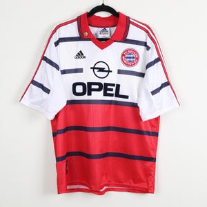 May include: A vintage FC Bayern Munich soccer jersey. The shirt is white with red and navy blue stripes and accents. The Opel logo is prominently displayed on the front, along with the Adidas logo and the team's crest.