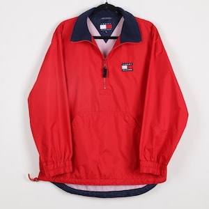 May include: A red Tommy Hilfiger windbreaker jacket with a navy blue collar and accents. The jacket features a half-zip closure, a front pocket, and the Tommy Hilfiger logo on the chest. The jacket is hanging on a black hanger.