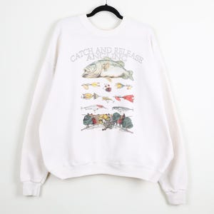 May include: White sweatshirt with a graphic featuring a fish, fishing lures, and the words "Catch and Release Angling." The design includes a detailed illustration of a fish and various fishing lures in multiple colors. The sweatshirt has long sleeves and a crew neck.