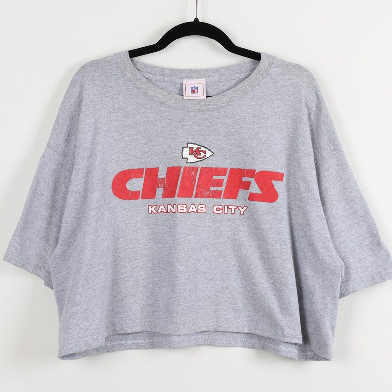 May include: A gray, short-sleeved t-shirt with the word "CHIEFS" in red block letters and the words "KANSAS CITY" underneath. The shirt also features the NFL logo.