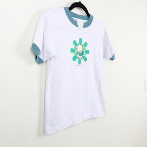 Puede incluir: Una camiseta gris con un ribete azul. La camiseta presenta un conejito de dibujos animados que lleva una camisa azul y est&aacute; de pie en una flor verde y amarilla. El texto "The Adventures of" est&aacute; impreso debajo del conejito.
