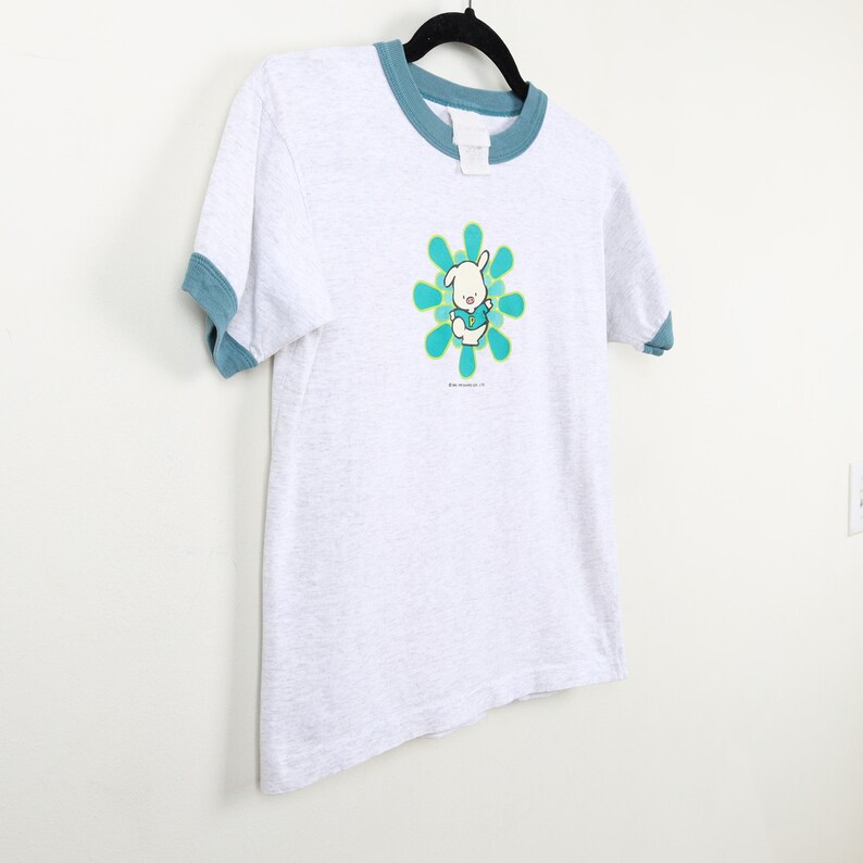 Puede incluir: Una camiseta gris con un ribete azul. La camiseta presenta un conejito de dibujos animados que lleva una camisa azul y est&aacute; de pie en una flor verde y amarilla. El texto "The Adventures of" est&aacute; impreso debajo del conejito.