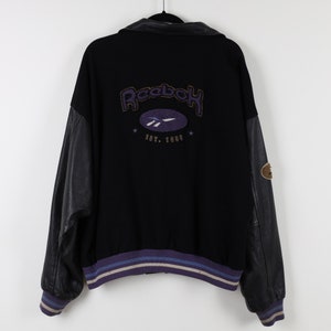 May include: Black Reebok varsity jacket with leather sleeves and a black collar. The back features the Reebok logo in purple and gold embroidery, with the text "EST. 1985". The jacket has a striped hem in purple, blue, and white.