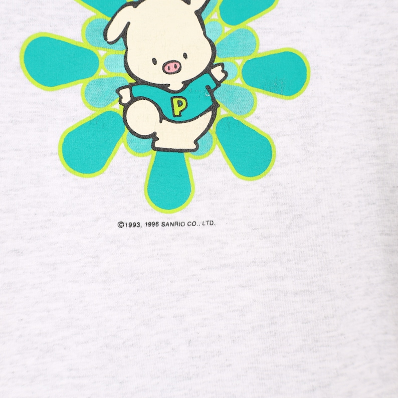 Puede incluir: Una camiseta gris claro con un cerdo de dibujos animados que lleva una camisa azul con una "P" blanca. El cerdo est&aacute; rodeado por un dise&ntilde;o circular turquesa y verde. El texto "&copy;1993, 1996 SANRIO CO., LTD." est&aacute; impreso en la parte inferior de la camiseta.