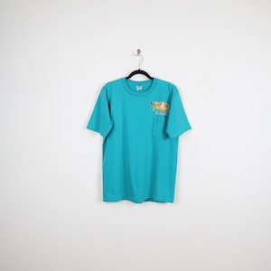 Puede incluir: Camiseta turquesa con un bolsillo en el pecho izquierdo. La camiseta tiene cuello redondo y mangas cortas. Un gr&aacute;fico colorido con texto est&aacute; impreso en el bolsillo. La camiseta est&aacute; colgada de una percha negra sobre un fondo blanco.
