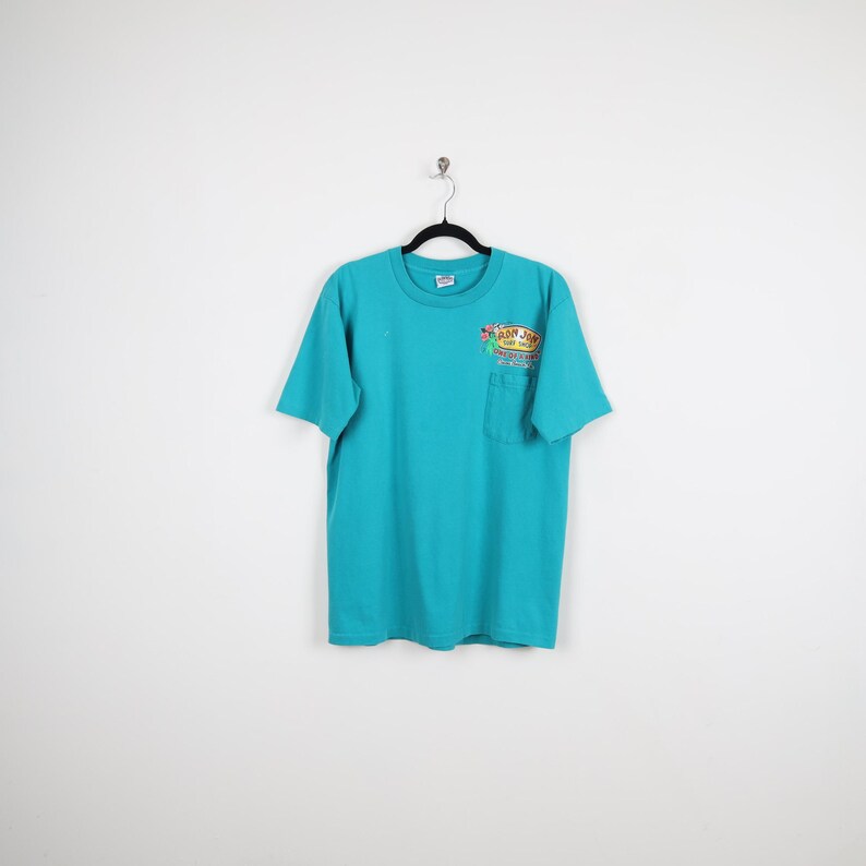 Puede incluir: Camiseta turquesa con un bolsillo en el pecho izquierdo. La camiseta tiene cuello redondo y mangas cortas. Un gr&aacute;fico colorido con texto est&aacute; impreso en el bolsillo. La camiseta est&aacute; colgada de una percha negra sobre un fondo blanco.
