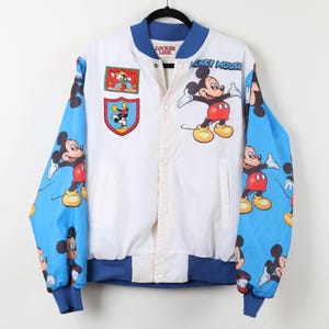 May include: White and blue Mickey Mouse bomber jacket. The jacket features a white body with blue sleeves patterned with Mickey Mouse. Mickey Mouse graphics are also on the front. The jacket has a blue collar, cuffs, and hem. The text "Locker Line" is visible.