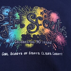 May include: A navy blue t-shirt with a colorful graphic of fireworks and the text "Girl Scouts 1912-2012 Celebrating 90 years! Girl Scouts of Santa Clara County".