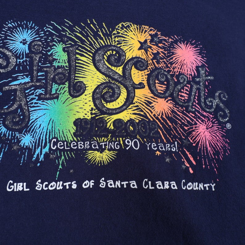 May include: A navy blue t-shirt with a colorful graphic of fireworks and the text "Girl Scouts 1912-2012 Celebrating 90 years! Girl Scouts of Santa Clara County".