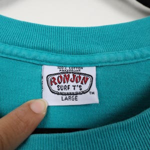 Puede incluir: Primer plano de un cuello de camiseta turquesa con una etiqueta blanca. La etiqueta dice "RONJON SURF T'S" en un dise&ntilde;o rojo y negro, con la palabra "LARGE" debajo. La camiseta es 100% algod&oacute;n y est&aacute; preencogida.