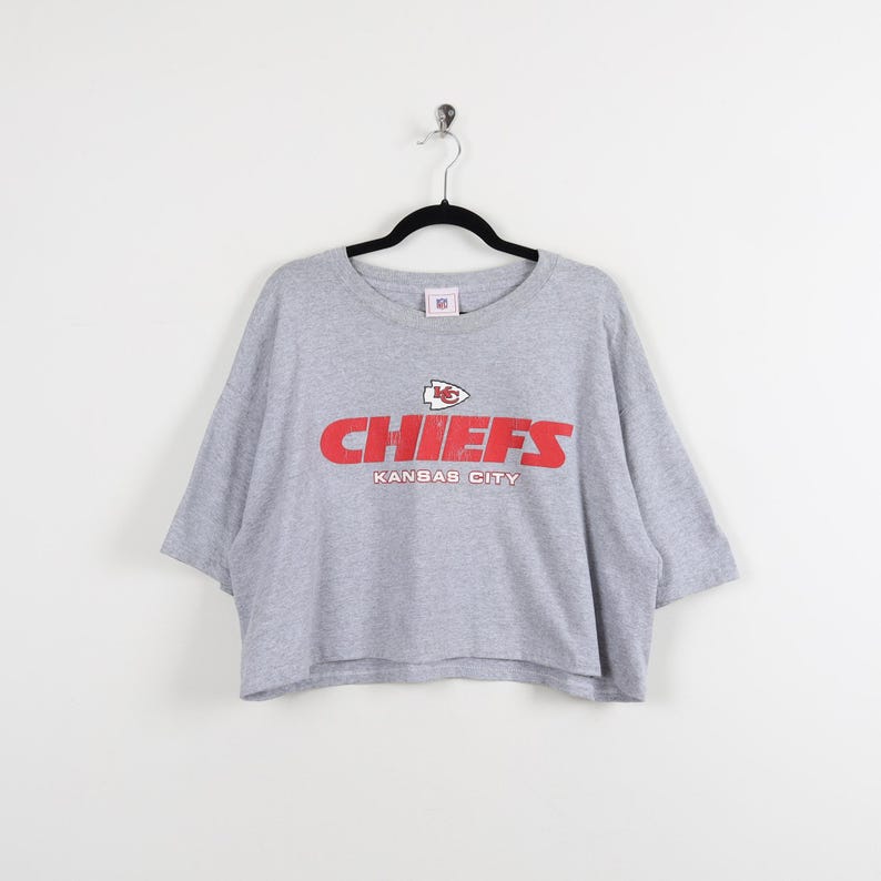 May include: A gray Chiefs t-shirt with a crew neck and short sleeves. The red text "CHIEFS" and "KANSAS CITY" are printed on the front. The shirt is hanging on a black hanger against a white background.