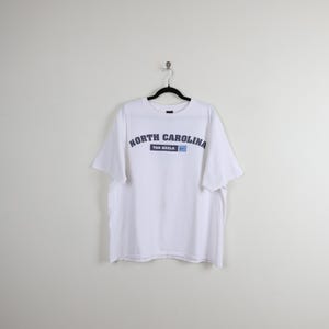 May include: White t-shirt featuring the text "NORTH CAROLINA" in navy blue arched lettering. Below the text is "TAR HEELS" in navy blue, with a small blue Nike swoosh logo. The shirt is suspended from a black hanger.