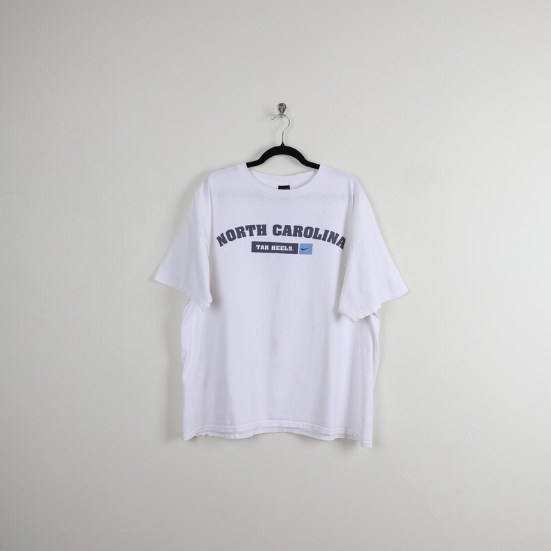 May include: White t-shirt featuring the text "NORTH CAROLINA" in navy blue arched lettering. Below the text is "TAR HEELS" in navy blue, with a small blue Nike swoosh logo. The shirt is suspended from a black hanger.