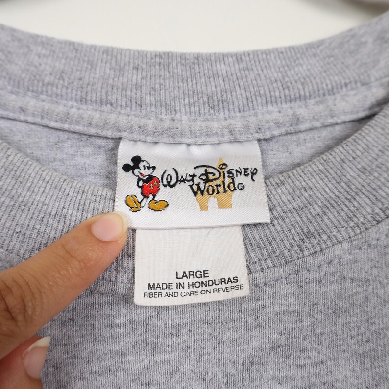 Pode incluir: Close-up de uma gola de camiseta cinza com uma etiqueta da Walt Disney World. A etiqueta apresenta um gr&aacute;fico do Mickey Mouse e o texto "Walt Disney World". Abaixo, o texto "LARGE" e "MADE IN HONDURAS".
