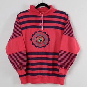 May include: A red and purple striped sweatshirt with a high collar and a drawstring. The sweatshirt features a circular emblem with text that reads "THE WORLD ACCREDITED ROYAL CLUB TENNIS". The sleeves and body have horizontal stripes.
