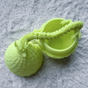 May include: A light green, 3D-printed dragon-shaped container with a lid. The container is round with a textured, scaly design. The lid is open, revealing the inside. A dragon's body and tail form the handle.