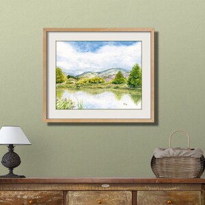 May include: A framed watercolor painting of a lake scene with green hills and trees in the background. The sky is blue with white clouds. The lake is reflecting the sky and trees.