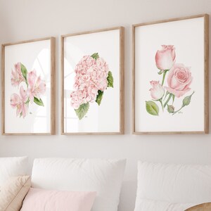 May include: Three framed watercolor paintings of pink flowers. The paintings are hung on a white wall above a white couch with pink throw pillows.