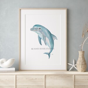 Op de afbeelding: Een aquarel schilderij van een blauwe dolfijn die uit het water springt. De dolfijn kijkt naar rechts en heeft een witte buik. Het schilderij is op een witte achtergrond en heeft de tekst "&copy; JAMES RIVER STUDIOS" in de linkerbenedenhoek.