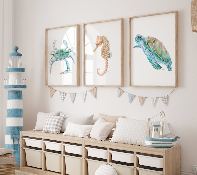 Op de afbeelding: Drie ingelijste aquarellen van zeedieren: een blauwe krab, een zeepaardje en een zeeschildpad. De schilderijen hangen aan een witte muur boven een houten bank met kussens en opbergbakken.