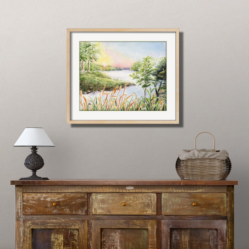 May include: A framed watercolor painting of a serene landscape. The artwork depicts a river scene with trees, reeds, and a colorful sunset. The frame is a light wood color, and the painting is displayed above a wooden cabinet.