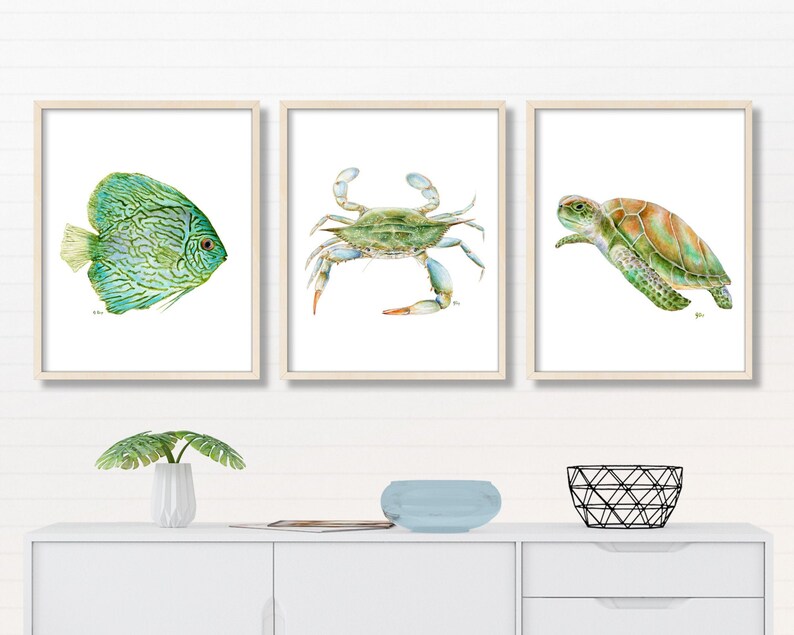May include: Set of three framed watercolor prints featuring a blue and green fish, a blue crab, and a green sea turtle. The prints are displayed on a white shelf with a small plant and a decorative bowl.