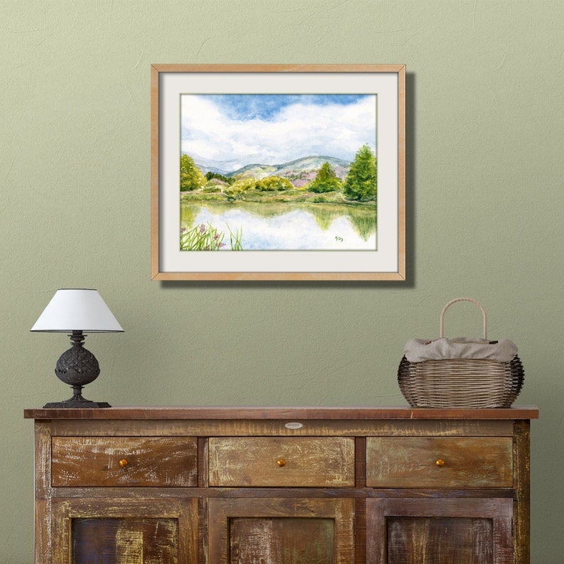 May include: A framed watercolor painting of a lake scene with green hills and trees in the background. The sky is blue with white clouds. The lake is reflecting the sky and trees.