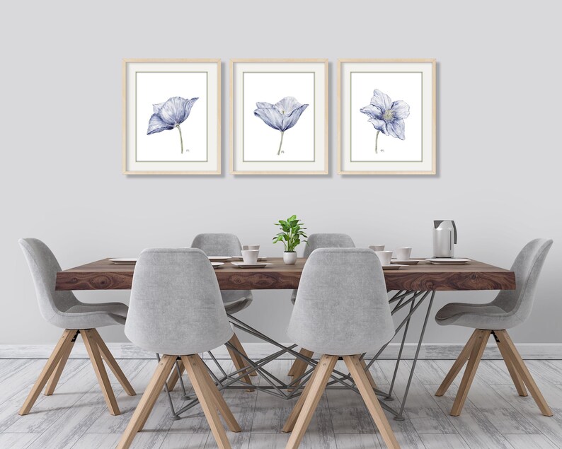 Puede incluir: Tres acuarelas enmarcadas de flores azules con centros blancos. Las pinturas est&aacute;n colgadas en una pared blanca sobre una mesa de comedor de madera con cuatro sillas.