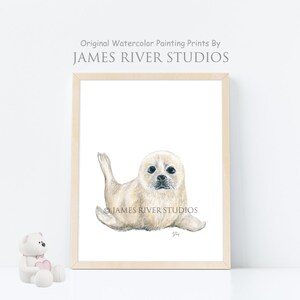 May include: A watercolor painting of a white baby seal with black eyes and a black nose. The seal is lying on its belly with its flippers outstretched. The painting is framed in a light wood frame.
