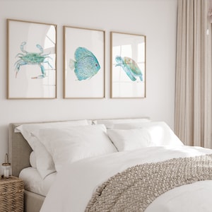 May include: Three framed watercolor paintings of sea life. The paintings are of a blue crab, a turquoise fish, and a green sea turtle. The paintings are hung on a white wall above a white bed with a beige headboard.