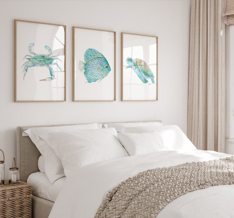 May include: Three framed watercolor paintings of sea life. The paintings are of a blue crab, a turquoise fish, and a green sea turtle. The paintings are hung on a white wall above a white bed with a beige headboard.