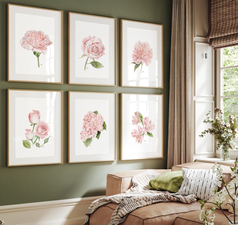 May include: Six framed watercolour paintings of pink flowers, including roses, peonies, and hydrangeas, displayed on a green wall above a light brown sofa with a striped throw blanket and cushions.