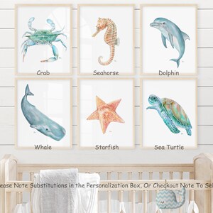 Op de afbeelding: Een set van zes aquarel illustraties van zeedieren, waaronder een krab, een zeepaardje, een dolfijn, een walvis, een zeester en een zeeschildpad. Elke illustratie is ingelijst in een licht houten lijst en wordt weergegeven op een witte muur.