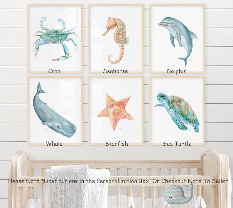 Op de afbeelding: Een set van zes aquarel illustraties van zeedieren, waaronder een krab, een zeepaardje, een dolfijn, een walvis, een zeester en een zeeschildpad. Elke illustratie is ingelijst in een licht houten lijst en wordt weergegeven op een witte muur.
