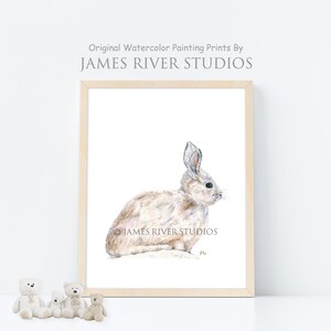 May include: A watercolor painting of a white rabbit sitting on a white background. The painting is framed in a light wood frame. The artist's signature is visible in the bottom right corner of the painting.
