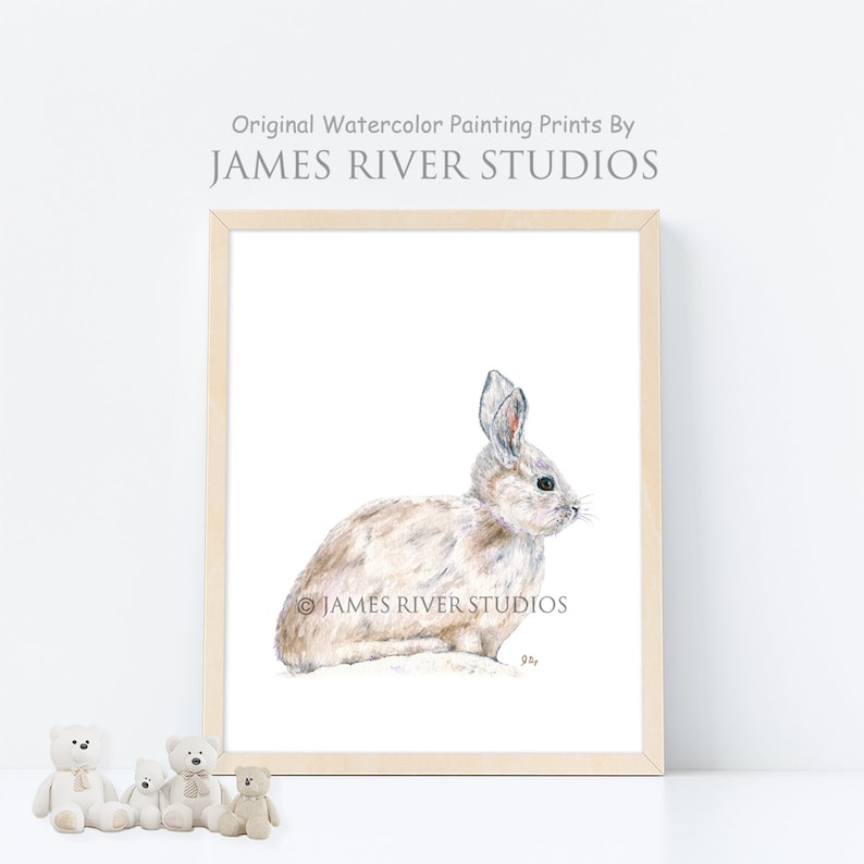 May include: A watercolor painting of a white rabbit sitting on a white background. The painting is framed in a light wood frame. The artist's signature is visible in the bottom right corner of the painting.