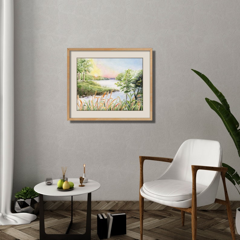 May include: A framed watercolor painting of a serene landscape featuring a river or lake, trees, and a colorful sky. The artwork is displayed on a gray wall above a white chair and a small table with decor.