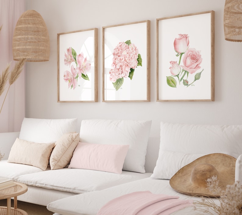 May include: Three framed watercolor paintings of pink flowers. The paintings are hung on a white wall above a white couch with pink throw pillows.