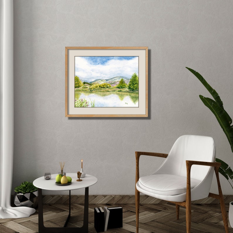 May include: A framed watercolor painting of a lake scene with green hills and trees in the background. The sky is blue with white clouds. The lake is reflecting the sky and hills.