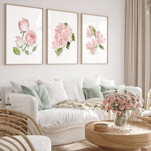 May include: Three framed watercolor prints of pink flowers. The prints are hung on a white wall above a white couch with green throw pillows.