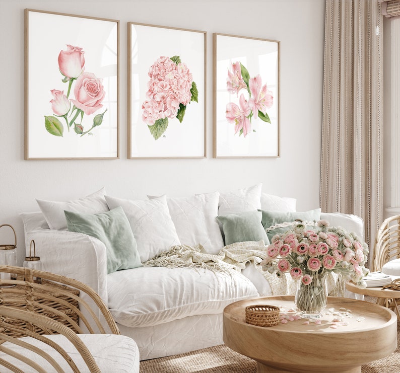 May include: Three framed watercolor prints of pink flowers. The prints are hung on a white wall above a white couch with green throw pillows.