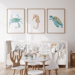 Op de afbeelding: Drie ingelijste aquarel illustraties van zeedieren: een blauwe krab, een zeepaardje en een zeeschildpad. De lijsten zijn van lichtbruin hout en de illustraties zijn op wit papier. De lijsten hangen aan een witte muur boven een wit wiegje met een wit-zwart gestreept kussen en een wit wolkenvormig kussen.