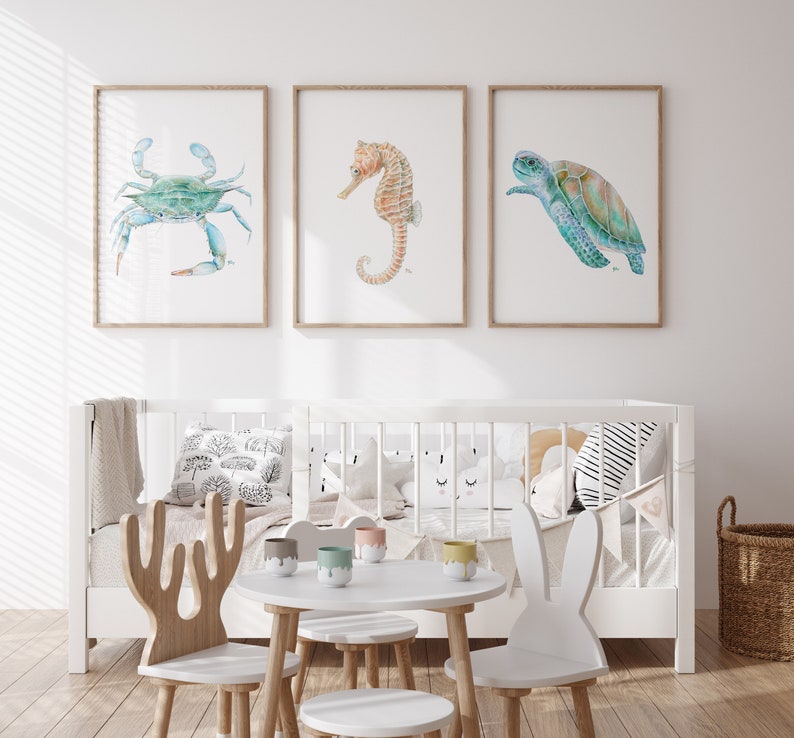 Op de afbeelding: Drie ingelijste aquarel illustraties van zeedieren: een blauwe krab, een zeepaardje en een zeeschildpad. De lijsten zijn van lichtbruin hout en de illustraties zijn op wit papier. De lijsten hangen aan een witte muur boven een wit wiegje met een wit-zwart gestreept kussen en een wit wolkenvormig kussen.