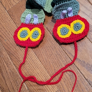 May include: A handmade crochet caterpillar toy in vibrant colors. The caterpillar features a red head with yellow and blue eyes, a green and blue body, and red string antennae. The toy is crafted with a textured stitch.