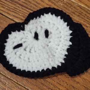 May include: A handmade, crocheted coaster featuring a cartoon dog face. The coaster has a white center with black eyes, a nose, and a black outline. The coaster is round and sits on a wooden surface.