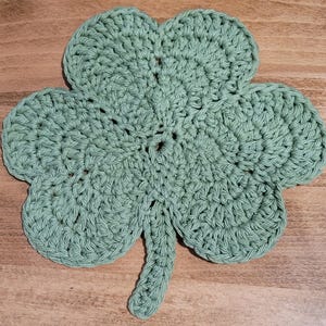 May include: A crocheted shamrock-shaped coaster in a light green color. The coaster is made of yarn and has a three-leaf clover design.